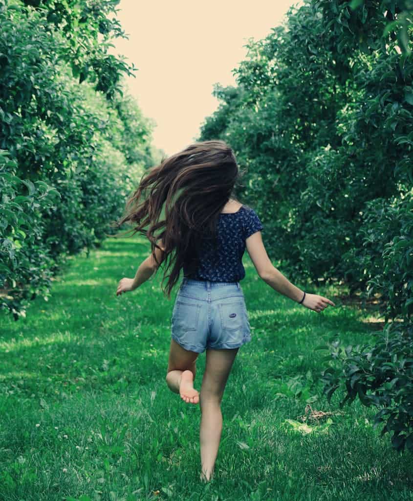 A girl joyfully runs through a sunlit orchard, enjoying nature and freedom.