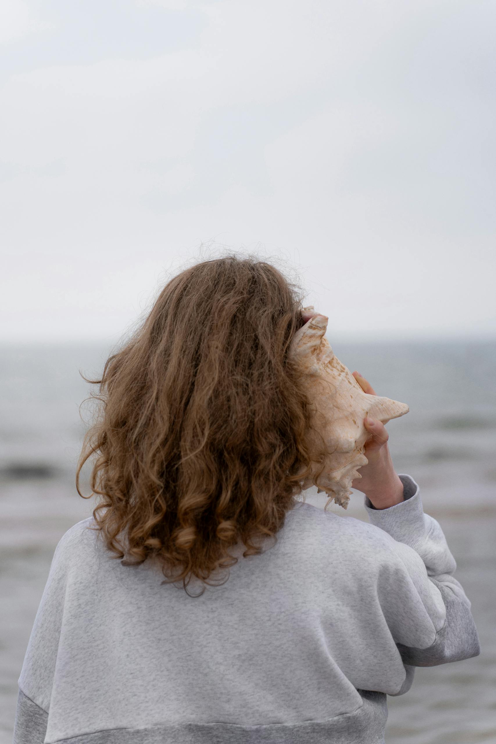 A young woman listens to a large seashell on a cloudy beach day.