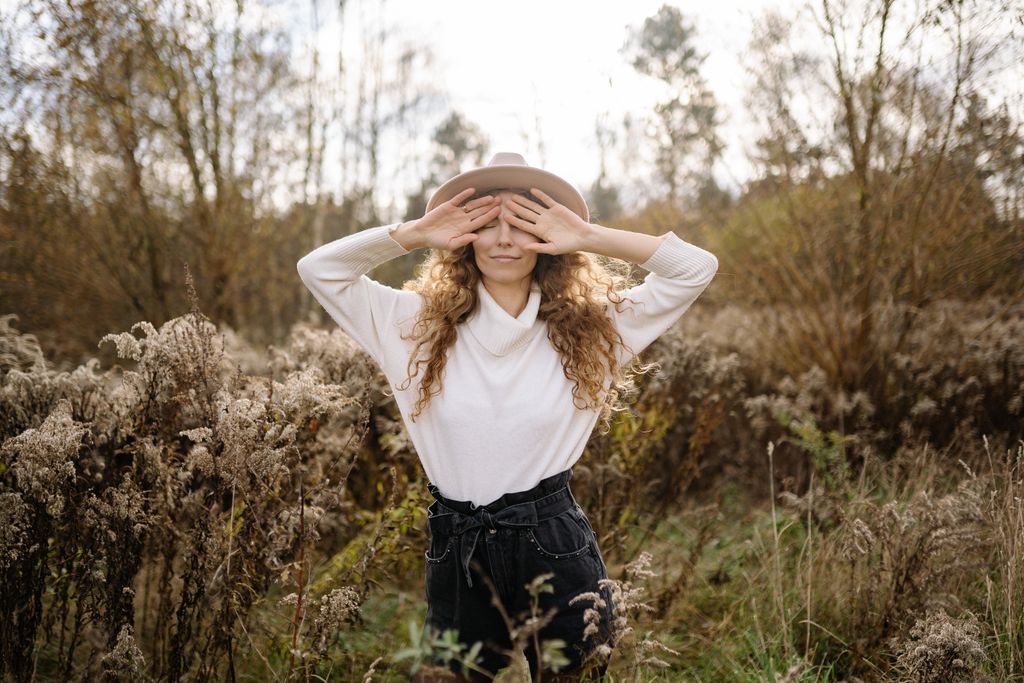 Woman in a hat playfully covering her eyes in an autumn field, showcasing nature and fashion.